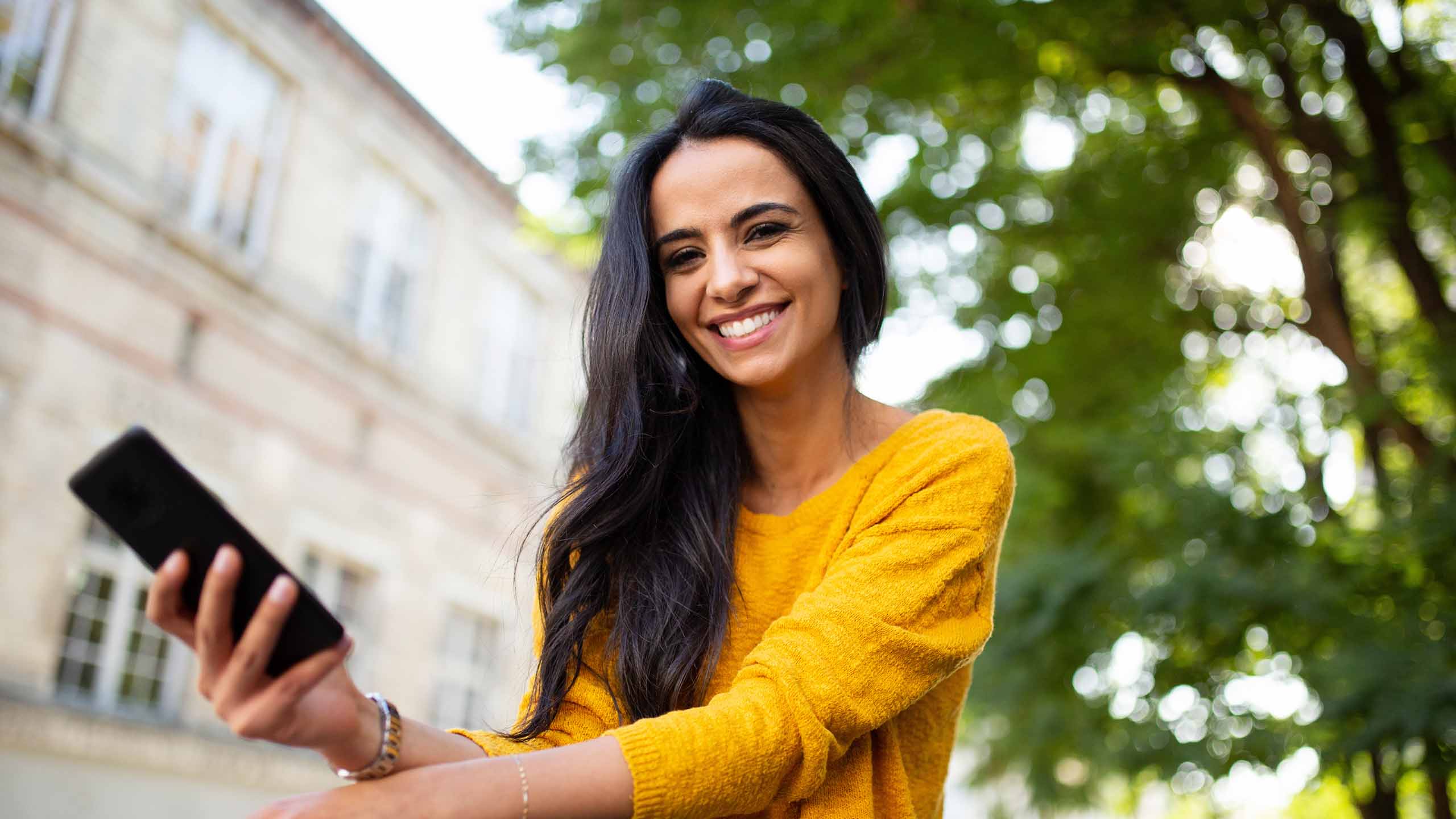 young woman smiling with mobile phone in hand outside