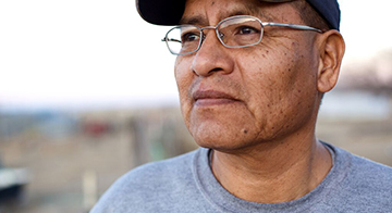 Man of colour wearing spectacles, a baseball cap and a grey t shirt looks into the middle distance