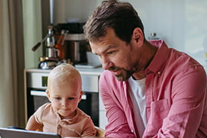 Man sitting at his kitchen table showing the screen of his tablet to young child who is in a highchair.