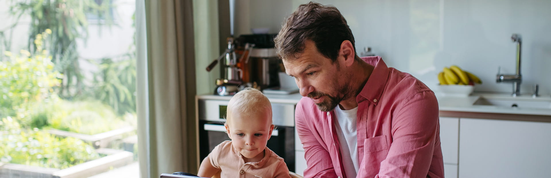 Man sitting at his kitchen table showing the screen of his tablet to young child who is in a highchair.