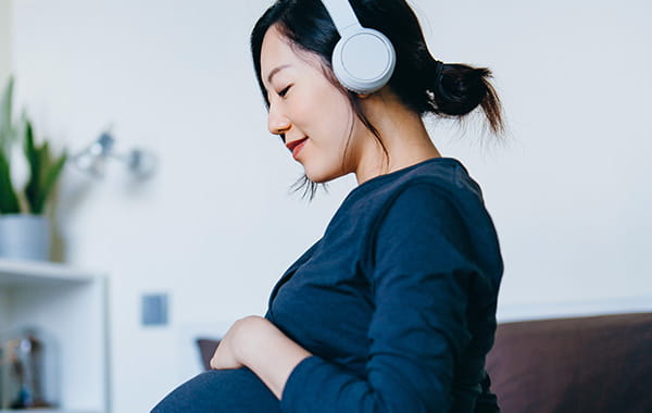 Pregnant woman sitting comfortably with headphones on and smiling as she holds her hand across her belly.