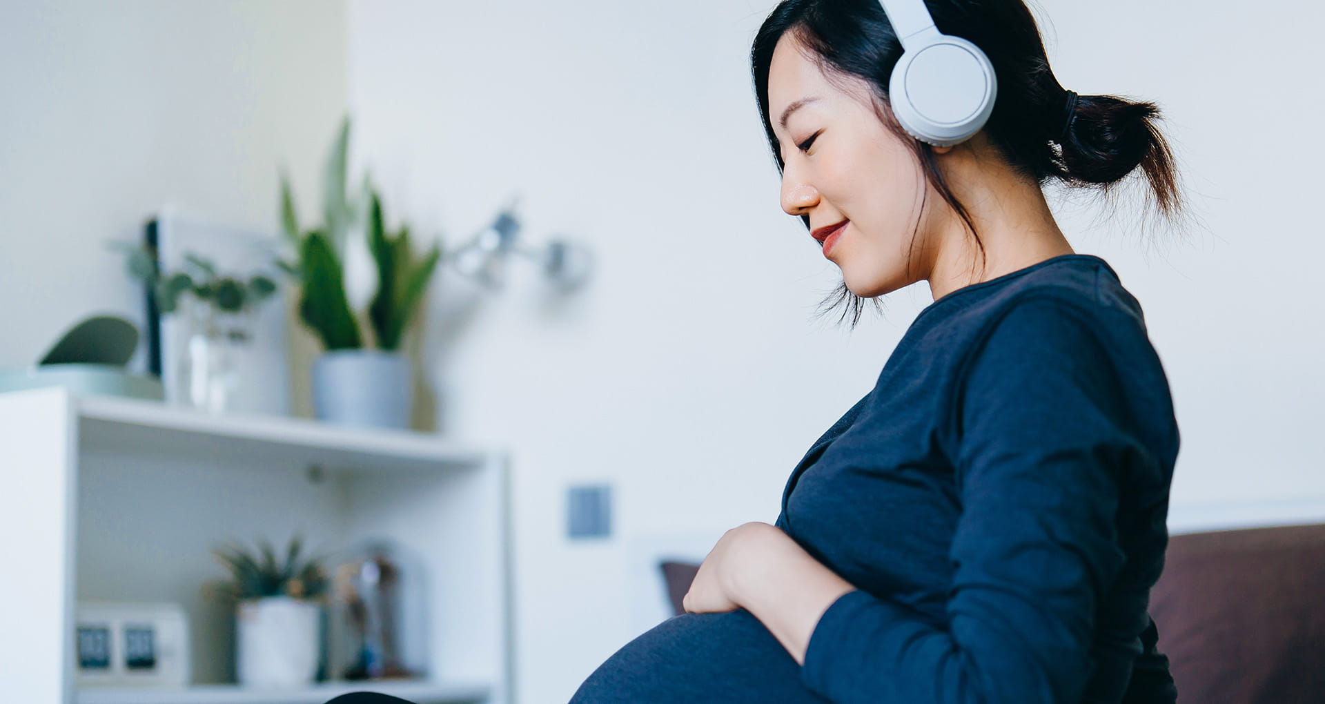Pregnant woman sitting comfortably with headphones on and smiling as she holds her hand across her belly.