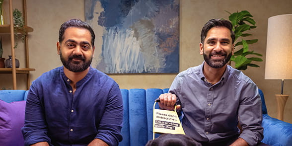 Dr Karan Rajan sitting next to Dr Amit Patel on a blue sofa in the studio, with Amit's dog Quark at his feet  