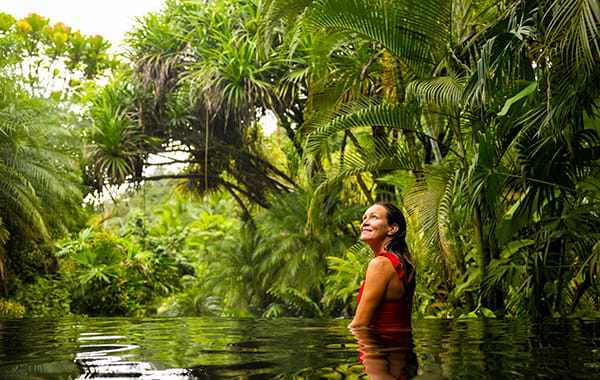 Woman standing in waist-height water in a rainforest environment and smiling while looking in wonder at the trees.