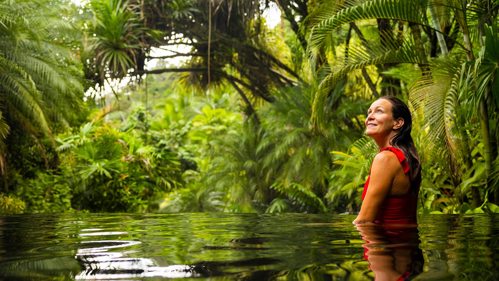 Woman standing in waist-height water in a rainforest environment and smiling while looking in wonder at the trees.