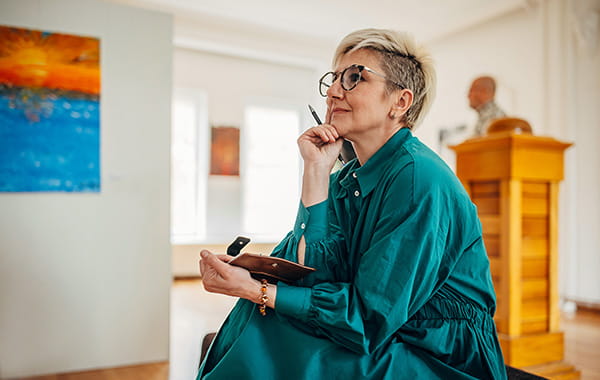 Woman wearing glasses and a turqoise dress, sitting attentively with a pen in her hand