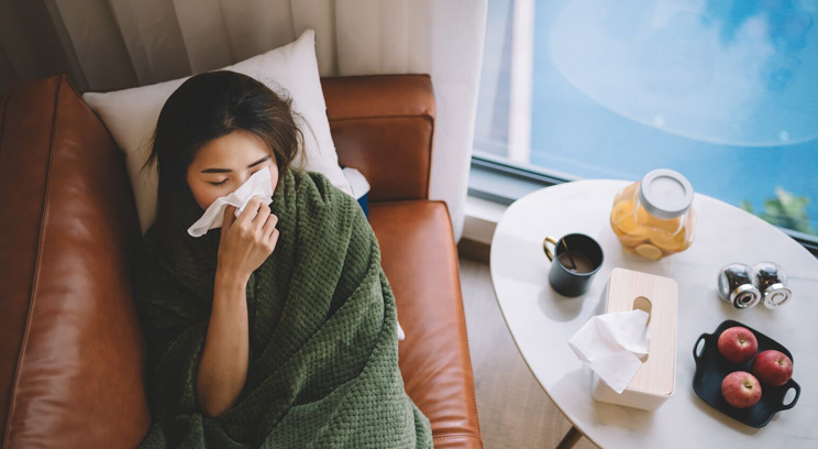 Young woman, wrapped in a blanket on the sofa, feeling unwell and holding a tissue to her nose.