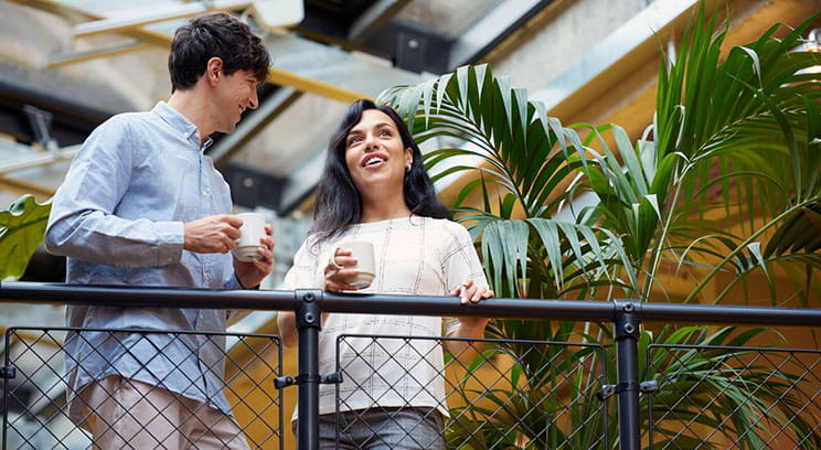 Two colleagues relaxed and chatting while standing on a balcony in the office and drinking coffee 