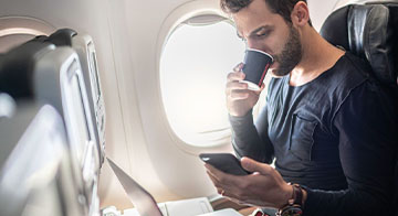 Man seated on plane drinking coffee and looking at his phone with a laptop also open on the tray in front of him 