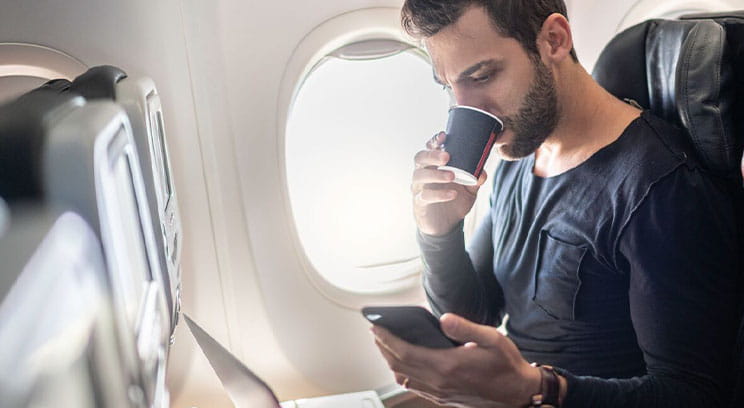 Man seated on plane drinking coffee and looking at his phone with a laptop also open on the tray in front of him 