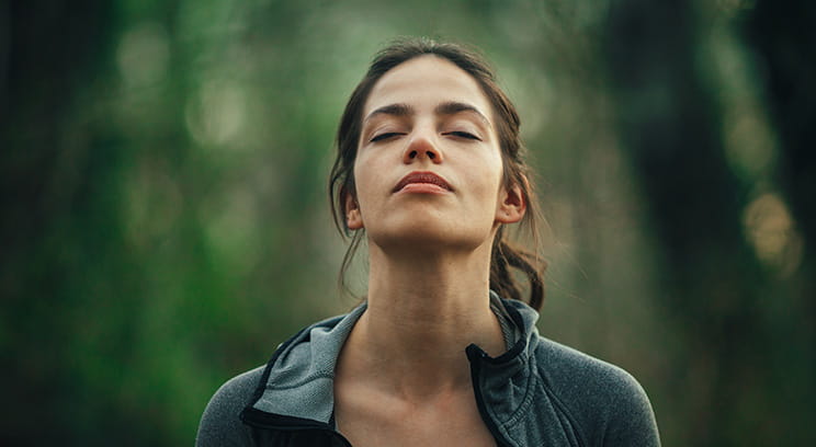 Young woman standing surrounded by trees in a forest, at peace with her eyes closed 