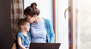Woman sitting at a window seat at home, working at laptop while embracing young child.