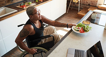 Woman of colour using wheelchair takes bowl of salad from kitchen counter