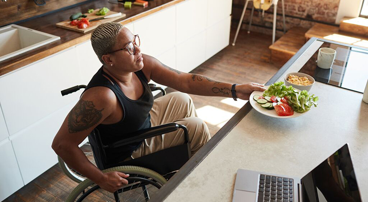Woman using wheelchair takes bowl of salad from kitchen counter