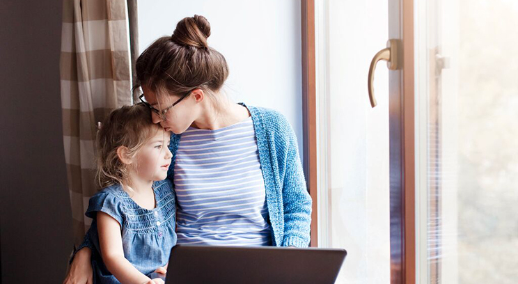 Woman sitting at a window seat at home, working at laptop while embracing young child.
