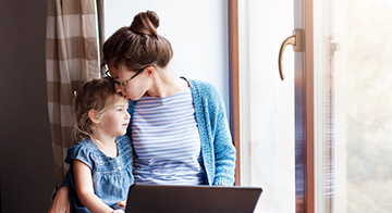Woman sitting at a window seat at home, working at laptop while embracing young child.