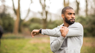 Man in grey hoodie stretching his arms outside in park with trees in the background before a run