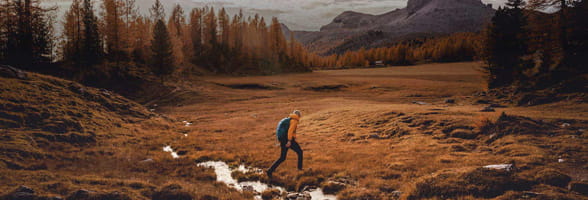 man crossing a river on a hike with mountains in the background