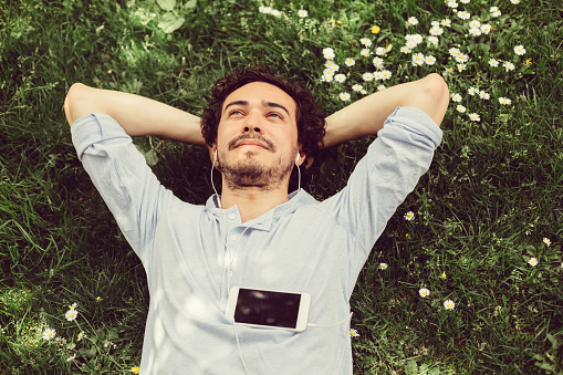 Man lying on grass with daisies, listening to media on mobile device