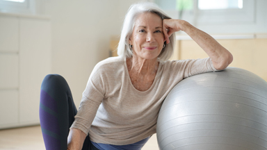 woman with grey hair smiling while sitting next to exercise ball